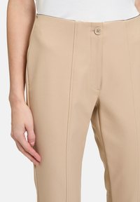 Béžové kalhoty na míru s hladkou texturou, disponující zapínáním na předním knoflíku a vertikálními švy pro strukturovaný střih.