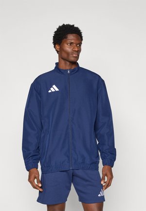 Navy blue athletic jacket with a high collar and full zip, featuring white Adidas logo accents. Made from a lightweight, smooth material.