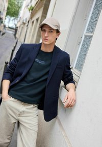 Blazer marine, t-shirt vert foncé avec du texte, pantalon beige clair et casquette gris clair. Tissu lisse et ajusté avec des détails sur les poches.