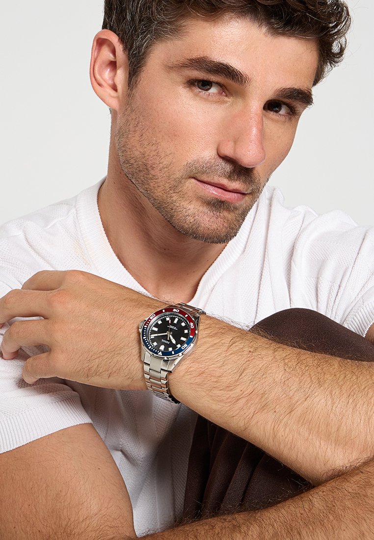 Reloj con esfera negra y pulsera de acero inoxidable plateado, bisel azul y rojo, números árabes y manecillas luminiscentes.