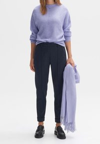 Žena v lila svetru, s tmavými kalhotami na míru, černými mokasínami, v jedné ruce drží odpovídající lila šálu s třásněmi.