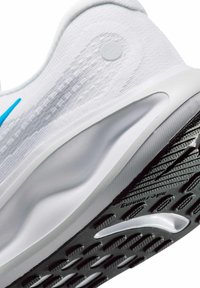 Witte sportieve schoen met een gestructureerde bovenkant, een gladde tussenzool en een zwarte rubberen buitenzool met een patroon voor grip. Blauwe accenten aan de zijkant.