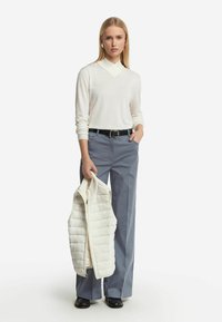 Cremefarbenes Langarmtop mit V-Ausschnitt, graue weite Hose und eine weiße gesteppte Weste, die in einer Hand gehalten wird. Ein schwarzes Gürtel und schwarze Schuhe vervollständigen das Outfit.