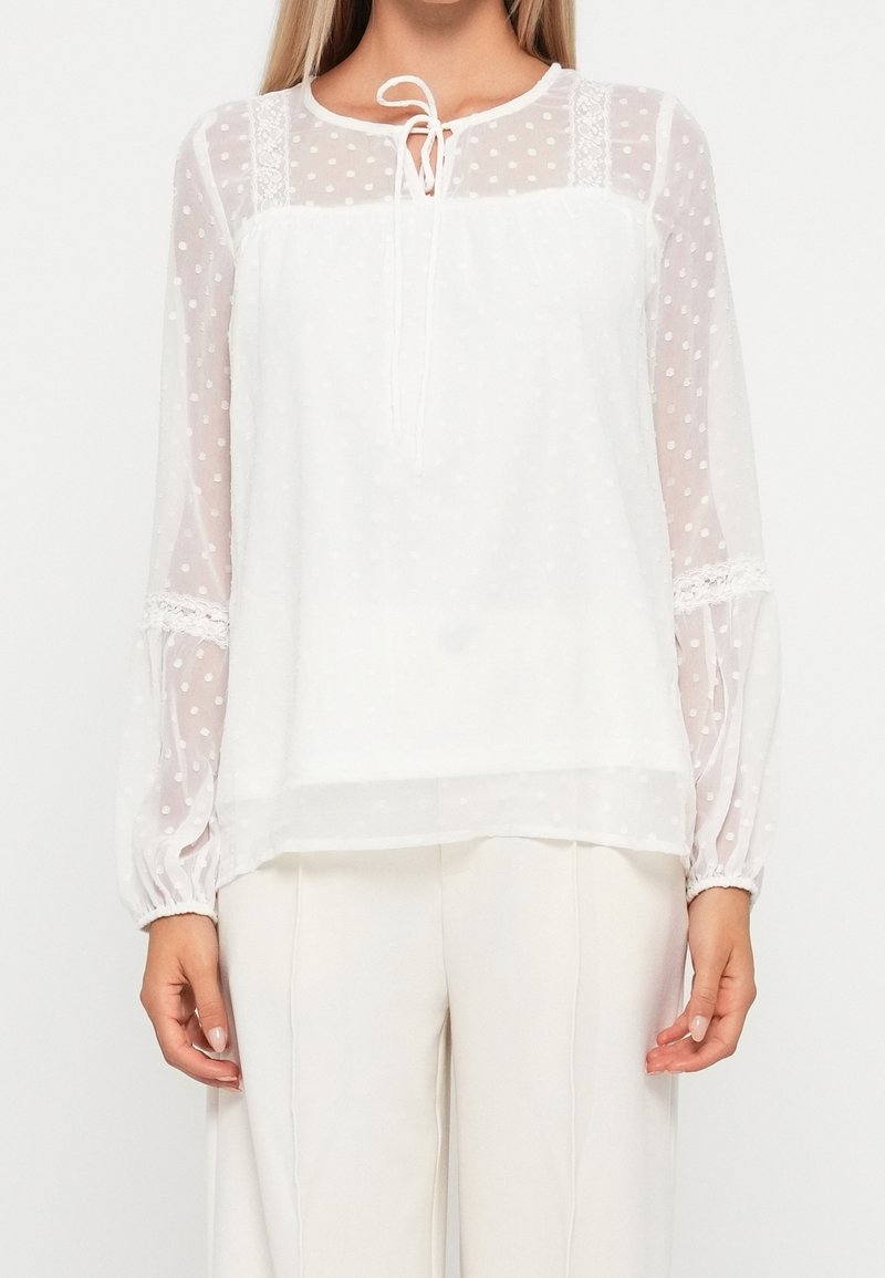 Blusa bianca con tessuto a pois semitrasparenti, maniche lunghe, dettagli in pizzo e un laccetto al collo. Taglio dritto, texture leggera.