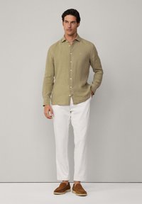 Camicia in lino verde chiaro con abbottonatura frontale, maniche lunghe e un logo discreto, abbinata a pantaloni bianchi e scarpe marroni.