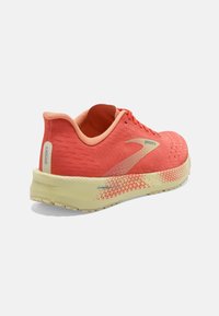 Chaussure de course Brooks en corail vif avec tige en maille texturée, col rembourré et semelle intermédiaire jaune présentant un motif de design géométrique.