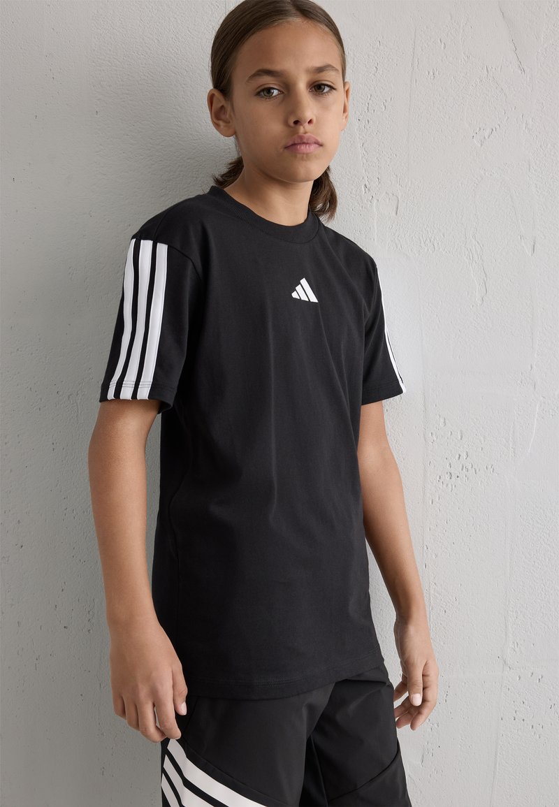 Sort bomulds T-shirt med hvidt Adidas-logo og stribede ærmer. Har en klassisk rund hals og afslappet pasform. Tekstureret væg baggrund.