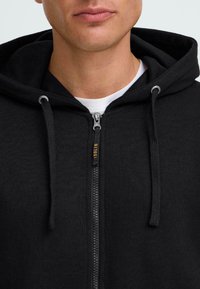 Hoodie zippé noir en tissu texturé, avec capuche à cordon de serrage et fermeture éclair pleine longueur. Superposé sur un T-shirt blanc en dessous.