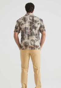 Chemise à manches courtes dans un motif abstrait beige et marron, avec une texture douce et un col, associée à un pantalon jaune clair.