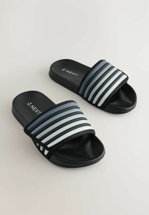 Mustad slaidid, millel on tekstureeritud tald ja lai rihm, millel on vahelduvad paksud triibud hallis ja valges. Brändi logo on nähtav rihmal.