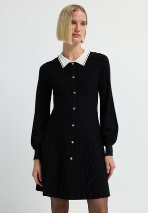 Zwarte jurk met lange mouwen, een witte kraag en knopen aan de voorkant. Plissé-ontwerp bij de rok en zilveren knoopaccenten overal.