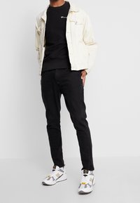 Uomo che indossa una giacca beige chiaro, una felpa nera Champion, jeans neri slim e sneakers bianche New Balance, in piedi contro uno sfondo bianco.