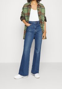 Femme portant un manteau à carreaux vert et marron, un haut blanc, un jean bleu évasé taille haute et des baskets blanches, se tenant avec une main dans la poche.