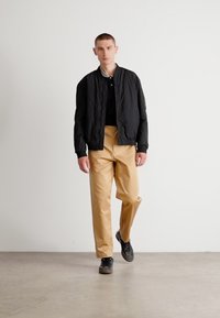 Black bomber jacket over a black polo, paired with beige trousers. Wearing dark shoes, on a light gray floor against a plain background.