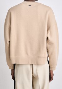 Beige oversized sweatshirt with a round neck, dropped shoulders, and ribbed cuffs; soft texture with a small logo tag at the back.