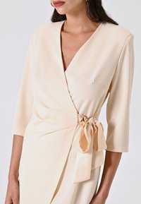 Wickelkleid aus leichtem beigen Kreppstoff, mit V-Ausschnitt, dreiviertellangen Ärmeln und einem dekorativen Bogen an der Taille mit einem goldenen Akzent.