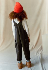 Sorte denim overalls med to frontlommer, hvid langærmet skjorte og rød ribbet strikbeanie. Brune sko og opsmøgede ærmer er synlige.