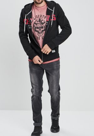 Zwarte hoodie met rits en rode letters, gedragen over een vervaagde roze t-shirt. Het model combineert het met grijze jeans en zwarte sportschoenen.