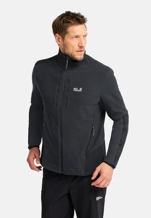 Mann trägt eine schwarze Jack Wolfskin Jacke mit Reißverschluss und schwarze Hose, schaut nach links vor einem schlichten weißen Hintergrund.