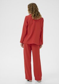 Femme aux cheveux bouclés portant un blazer rouge et un pantalon large, se tenant de dos sur un fond blanc uniforme.