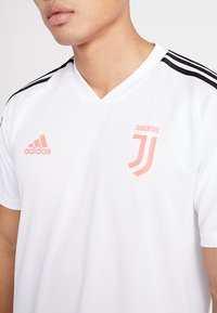 Vit Adidas-tröja med svarta accenter, med Juventus-logotypen i ljusrosa på bröstet och design med V-hals.