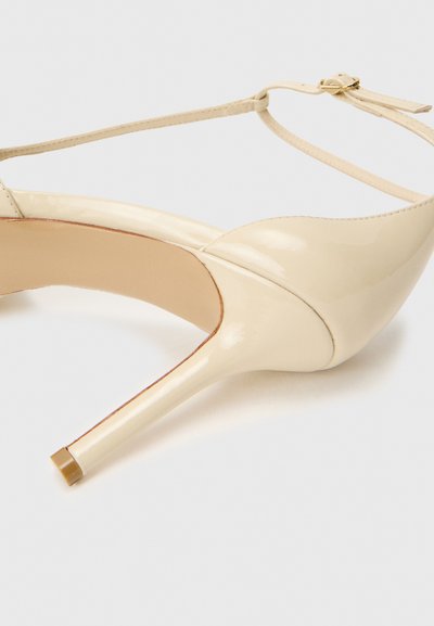Beige patent leather high-heeled shoe with a pointed toe, thin ankle strap, and a glossy finish. Features a slender stiletto heel.