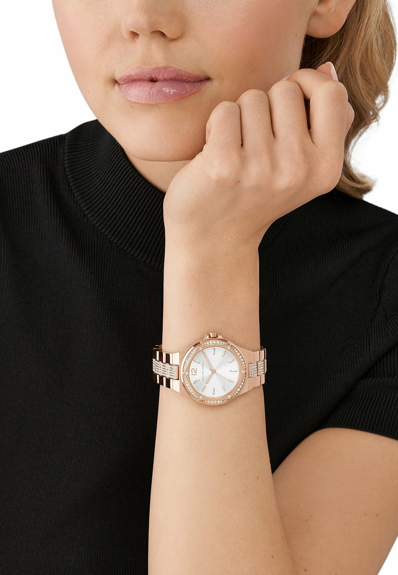Różowozłoty zegarek z okrągłą białą tarczą, dwu-tonową metalową bransoletą, błyszczącymi akcentami i znacznikami godzin. Noszony na nadgarstku w zestawie z czarną bluzką.