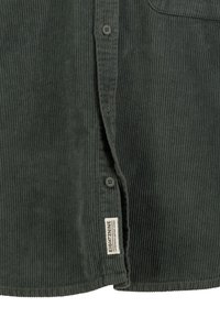 Dunkelgrünes Cordhemd mit Knöpfen vorne und einem weißen rechteckigen Stoffetikett mit der Aufschrift "EIGHT2NINE" nahe dem unteren Saum.