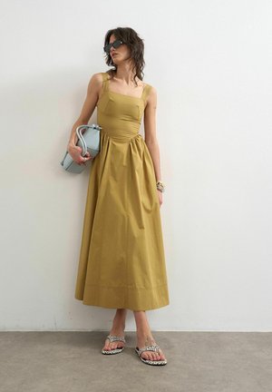 Femme en robe midi sans manches jaune moutarde, lunettes de soleil noires, tenant un sac à main bleu clair, portant des sandales à motifs, debout contre un mur blanc.