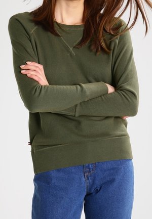 Woman with long brown hair wearing an olive green sweater and blue jeans, standing with arms crossed against a plain background.