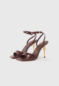 Mørkebrune højhælede sandaler med krokodillemønster, ankelremme og guld snoet metal stiletto hæle på en hvid baggrund.