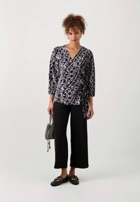 Haut enveloppant noir et blanc avec un motif géométrique, manches trois quarts, associé à un pantalon large noir et des chaussures plates. Sac à main gris.