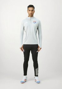 Sivi sportski top s dugim rukavima i četvrtastim patentnim zatvaračem, s logotipom. Uparen s crnim tajicama i šarenim nogometnim kopačkama.