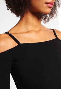 Donna con capelli ricci che indossa un top a coste nero con spalline sottili e clavicole in evidenza.