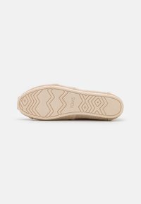 Bottine en toile beige à enfiler avec une semelle en caoutchouc texturée présentant des motifs en zigzag et le nom de la marque "TOMS" embossé au fond.