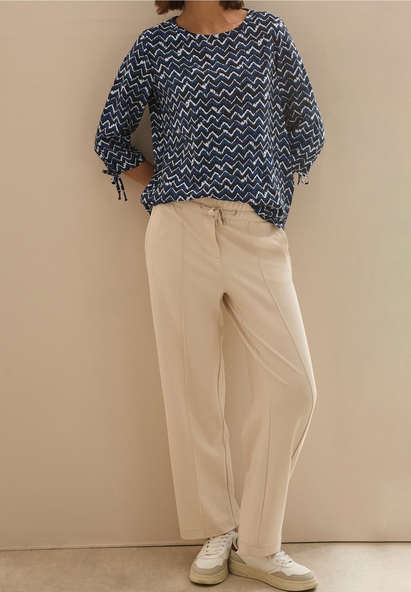 Personne portant un chemisier bleu à motif zigzag, un pantalon beige à cordon et des baskets blanches, appuyée de façon décontractée contre un mur uni.