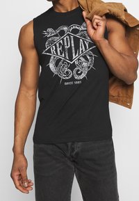 Camiseta sin mangas negra con un diseño gráfico que presenta serpientes y la palabra "REPLAY" en forma de diamante, fabricada en tejido de algodón.