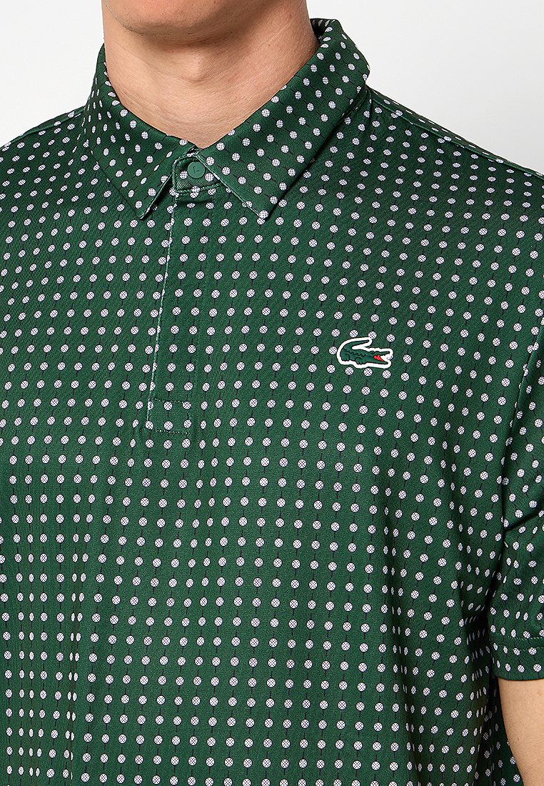 Hombre que lleva una camiseta polo verde oscuro con lunares blancos y un logo de cocodrilo en el pecho, abotonada en el cuello.