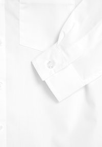 Poignet de chemise blanche à manches longues avec bouton, poche avant partielle et patte de boutonnage verticale sur une surface de tissu lisse.