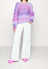 Stickad tröja i gradient av lila och rosa toner, breda ärmar, kombinerad med vita vida byxor och rosa träskor med ventilationshål.
