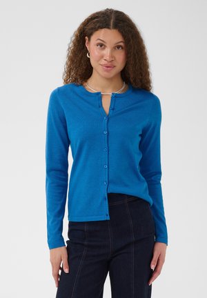Kvinde med krøllet hår iført en blå knap-op cardigan og mørke jeans, stående mod en ensfarvet lys baggrund.