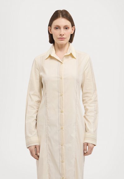 Rochie lungă de culoare crem, realizată dintr-un material neted, cu front butonat, guler ascuțit și detalii cu dungi verticale.