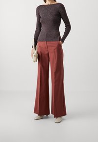 En kvinna står klädd i en mörk figurnära tröja och vida rostfärgade byxor, med en liten beige handväska i handen, mot en neutral bakgrund.