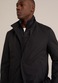 Chaqueta exterior negra con cuello alto, superpuesta a una chaqueta interior acolchada. Presenta botones visibles y bolsillos laterales. Textura suave.
