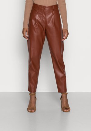 Kobieta ubrana w brązowe, wysokowalne spodnie ze sztucznej skóry oraz beżowe sandały na wysokim obcasie z paskiem wokół kostki, stojąca na białym tle.