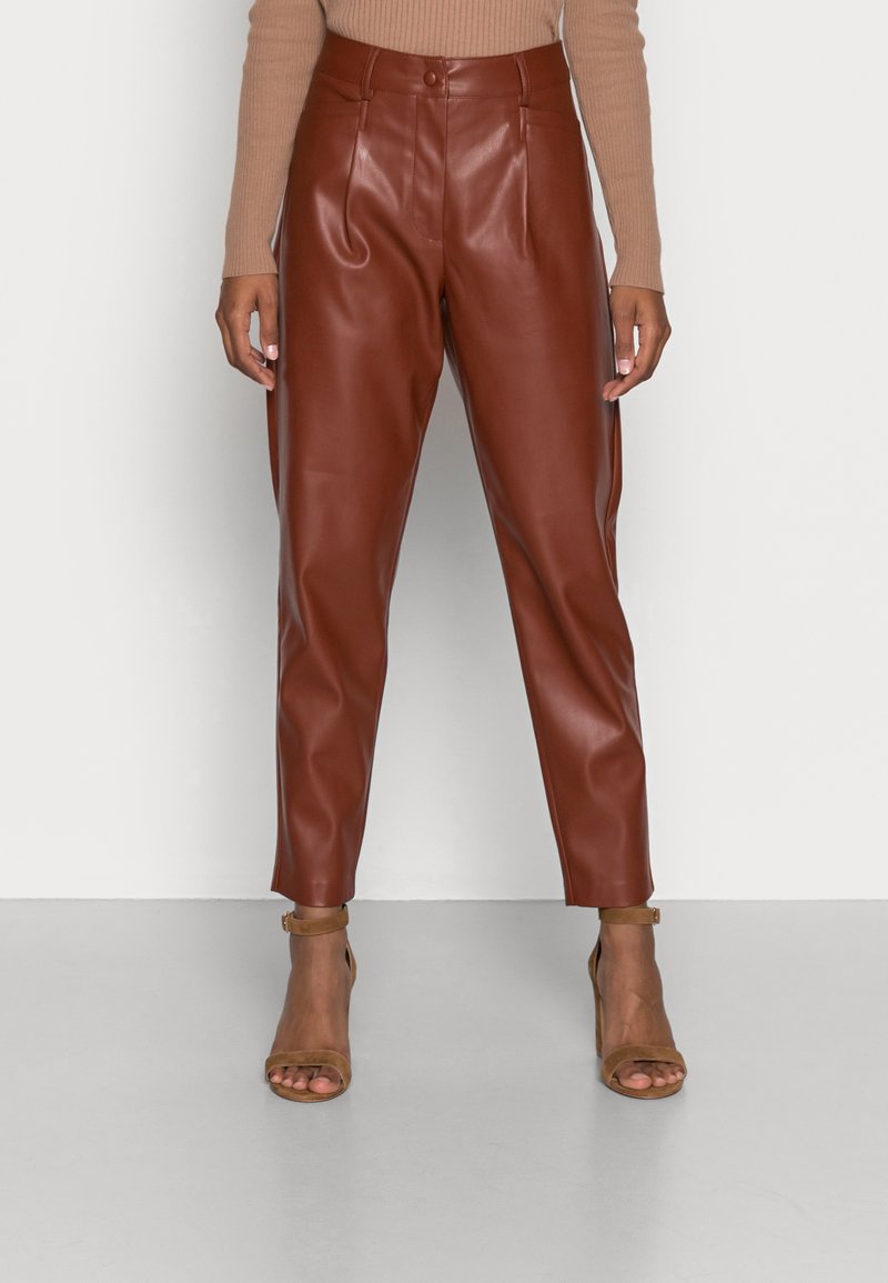 Femme portant un pantalon en similicuir marron taille haute et des sandales à talons hauts avec bride de cheville couleur beige, debout devant un fond blanc.