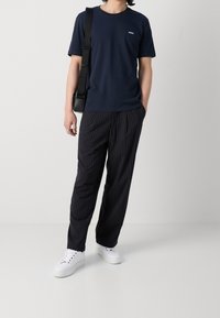 T-shirt in cotone blu navy con un piccolo logo bianco, abbinato a pantaloni neri a righe sottili e sneakers bianche, arricchito da una borsa a tracolla nera.