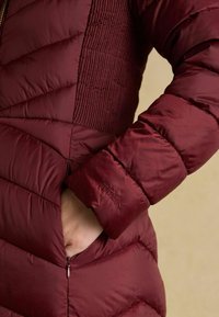 Burgundia värvi sulejope, millel on tikitud muster, varrukal on lukuga tasku ja tikitud logo, tehtud siledast ja läikivast kangast.