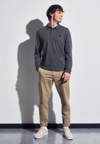 Muž v šedém dlouhém polo tričku, béžových kalhotách a bílých teniskách stojící s rukama v kapsách, dívající se doprava proti bílé zdi.