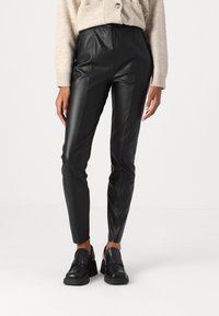 Zwarte faux leren broeken met een strakke uitstraling, hoge taille en opvallende details langs de zijkanten; gecombineerd met zwarte stevige loafers.